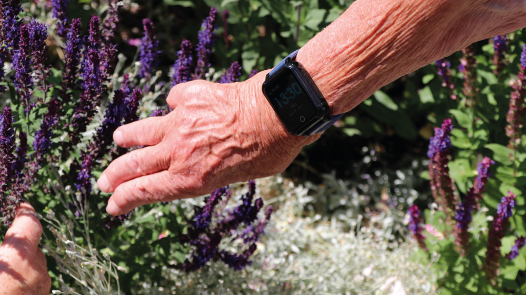 Sensorems smartklocka med trygghetslarm och gps ökar tryggheten för seniorer inomhus men också utomhus i trädgården på sommaren eller på resan. Bild på smarklocka på arm i trädgård